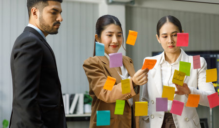 Millennial Asian Indian professional successful male businessman and female businesswomen colleagues in formal business suit standing pointing post it sticky note on glass board discussing together.の写真素材