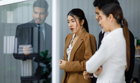 Millennial Asian professional businesswoman employee staff officer in formal business suit standing crossed arms with male female colleagues listening to Indian businessman discussing on glass board.の写真素材