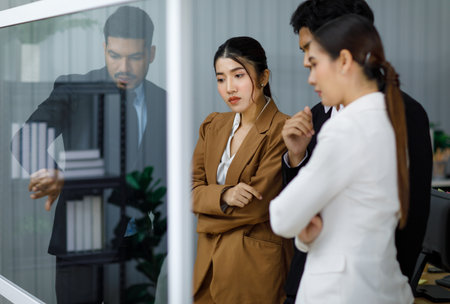 Millennial Asian professional businesswoman employee staff officer in formal business suit standing crossed arms with male female colleagues listening to Indian businessman discussing on glass board.の写真素材