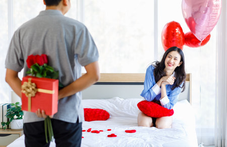 Millennial Asian young romantic lover couple unrecognizable male boyfriend holding red roses bouquet and wrapped gift box surprising female girlfriend on bed with heart shape balloon on valentine day.の写真素材