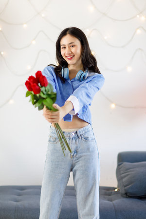 Millennial Asian young romantic cheerful female girlfriend standing in livingroom decorate with red heart shape balloon holding red roses bouquet smiling on anniversary valentine day celebrationの写真素材