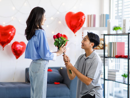Millennial Asian young romantic lover couple male boyfriend kneel down giving red roses bouquet surprising female girlfriend celebrating anniversary on valentine day festival in decorated living room.の写真素材