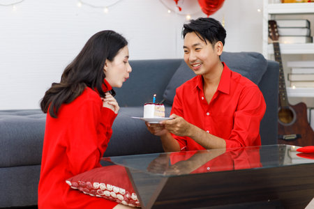 Millennial Asian young romantic lover couple male boyfriend in red outfit sitting on floor giving surprising female girlfriend with cake and candle celebrating happy birthday festival in living room.の写真素材