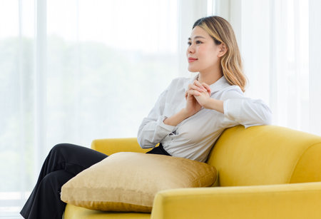 Asian professional successful cheerful happy thoughtful female businesswoman entrepreneur in formal outfit sitting alone on cozy sofa couch thinking business ideas solving problems in living room.の写真素材