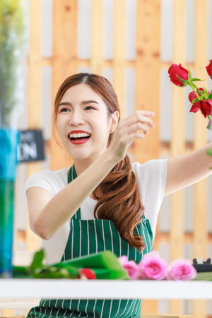 Asian professional successful female florist designer shop owner entrepreneur businesswoman sitting smiling holding decorating picking choosing red roses flowers bouquet in floral garden store studio.の写真素材