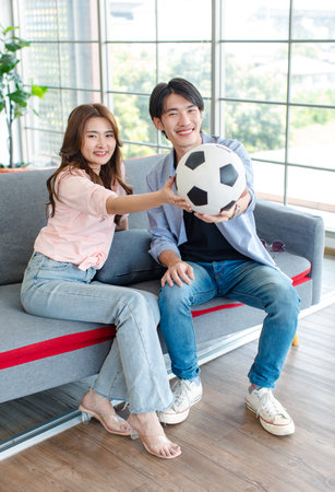 Asian cheerful excited male female football fan couple family sitting raising hands fists together on cozy sofa couch in living room holding ball cheering watching favorite team live on television.の写真素材