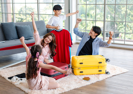 Asian little boy and girl helping father and mother preparing packing clothes and stuff into trolley luggages on living room carpet floor for traveling on holiday weekend vacation family road trip.の写真素材