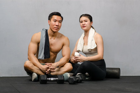 Asian strong young male muscular shirtless fitness model in sporty shorts and female athlete in sport bra sitting smiling taking break holding water bottle talking together in gym on gray background.の写真素材