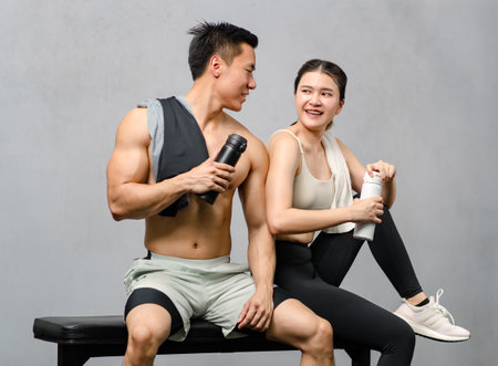 Asian strong young male muscular shirtless fitness model in sporty shorts and female athlete in sport bra sitting smiling taking break holding water bottle talking together in gym on gray background.の写真素材
