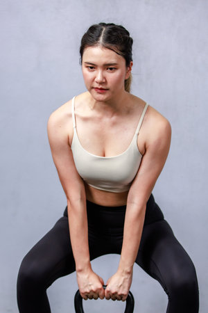 Asian strong beautiful young female muscular fitness model in sport bra and legging standing holding lifting metal kettlebell dumbbell showing biceps triceps muscle in gym on gray backgroundの写真素材