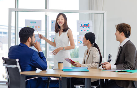 Millennial Asian professional successful businesswoman presenter in formal wears standing using presenting infographic paperworks documents on glass board in meeting room with  businessman groupの写真素材