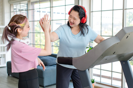 Asian old chubby retirement pensioner happy healthy female sport fitness woman in casual sportswear walking running on machine treadmill while young trainer holding raising hand high five in gymの写真素材