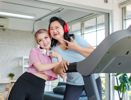 Asian happy healthy old chubby retirement pensioner mother smiling standing posing taking photo with young daughter wear headphones showing thumb up exercise on adjustable running pad in living room.の写真素材