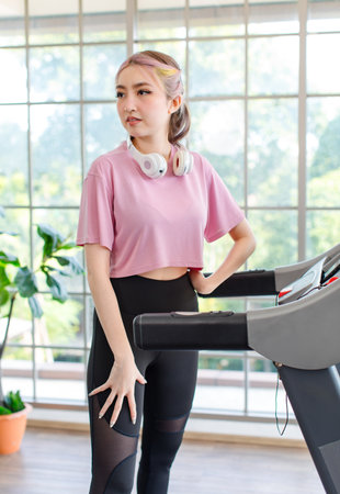 Portrait shot Asian unhappy young bored female sport fitness girl in casual sportswear with headphones standing holding hand on face on running machine treadmill looking at camera with frowning face.の写真素材