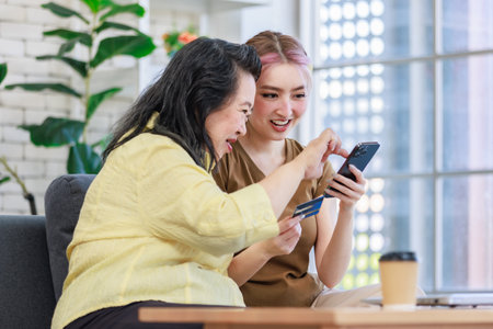 Asian young beautiful daughter sitting on cozy sofa holding touchscreen smartphone using credit card paying payment online via internet in living room at home with senior fat chubby pensioner mother.の写真素材