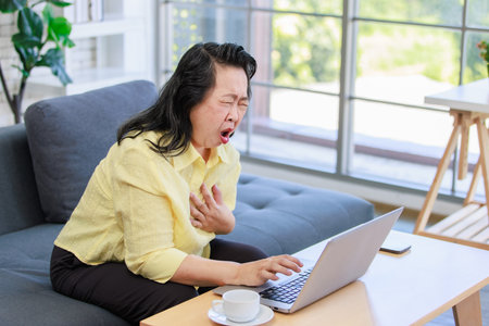 Asian old senior pensioner chubby fat housewife sitting on cozy sofa open mouth wide shocking holding hand on chest having heart attack while surfing browsing internet online via laptop computer.の写真素材