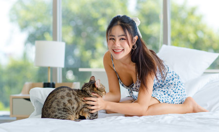 Asian young beautiful cheerful female teenager in sexy dress laying on white clean sheet bed with pillows smiling playing with cute domestic short hair pussycat pet in bedroom at home in morning.の写真素材
