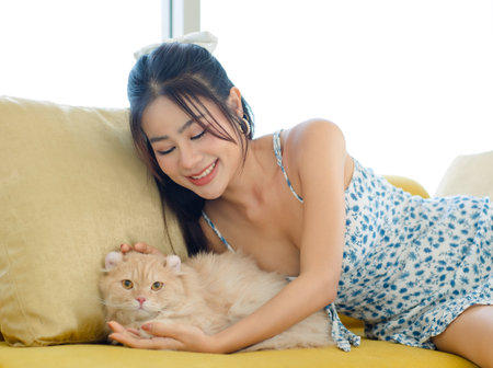 Asian young beautiful female teenager girl in sexy dress laying lying down on cozy sofa couch smiling cuddling hugging embracing relaxing little cute domestic orange long hair pussycat companion pet.の写真素材