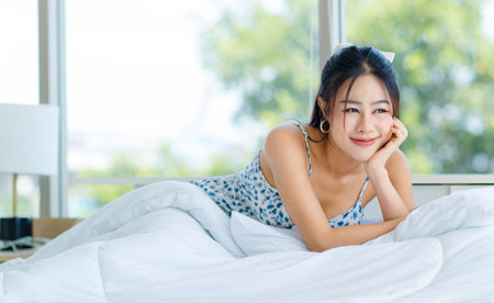 Asian young beautiful pretty cheerful female teenager girl in sexy pyjamas blue spotted dress sitting smiling waking up fresh on cozy white clean sheet bed under warm blanket in bedroom in morning.の写真素材