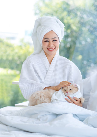 Asian young pretty sexy female teenager girl in bathrobe ready for relaxing spa on bed sitting smiling playing with cute little domestic orange long hair companion pet cat kitty in towel in bedroom.の写真素材