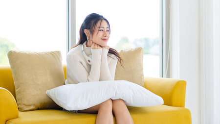 Asian young beautiful female teenager girl owner in turtleneck sweater wearing headphones listening to music sitting smiling on cozy sofa holding cute little orange long hair domestic cat on pillow.の写真素材