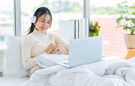 Asian pretty female teenager girl in turtleneck sweater wearing headphones listen to music from laptop on white clean sheet bed under blanket drinking coffee with cute domestic tabby short hair cat.の写真素材