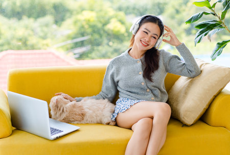 Asian young pretty cheerful female teenager girl in sweater wearing headphones listening to music from laptop sitting on cozy sofa couch smiling with little cute domestic long hair companion pet cat.の写真素材