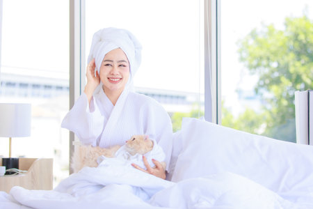 Asian young pretty sexy female teenager girl in bathrobe ready for relaxing spa on bed sitting smiling playing with cute little domestic orange long hair companion pet cat kitty in towel in bedroom.の写真素材
