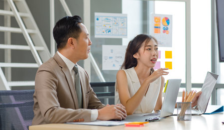 Asian professional successful male businessman manager mentor in formal business suit standing helping teaching supporting female businesswoman colleague sitting working with laptop notebook computer.の写真素材