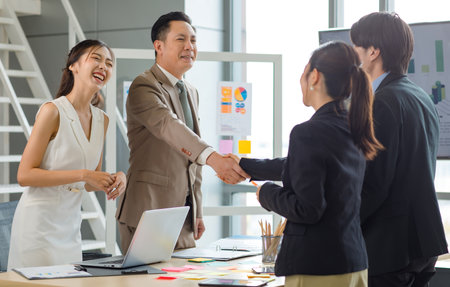 Asian professional successful businesswoman standing smiling shaking hands greeting with unrecognizable businessman in formal business suit while male female colleagues clapping hands celebrating.の写真素材