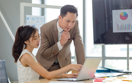 Asian professional successful male businessman manager mentor in formal business suit standing helping teaching supporting female businesswoman colleague sitting working with laptop notebook computer.の写真素材