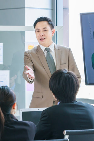 Asian professional successful male businessman lecturer speaker presenter in formal business suit standing at strategy data information monitor speaking to audiences in company office meeting room.の写真素材