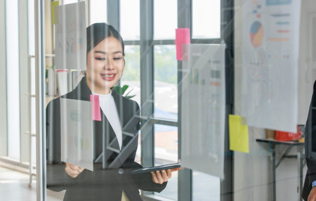 Asian professional successful female businesswoman lecturer presenter in formal business suit standing using marker pointing infographic paperwork document sticky note on glass board in meeting room.の写真素材