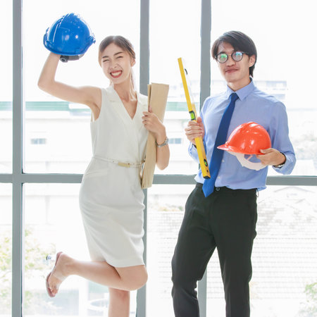 Asian professional successful male businessman and female businesswoman engineer architect smiling laughing hold safety hardhat and blueprint standing smiling celebrating together in company office.の写真素材