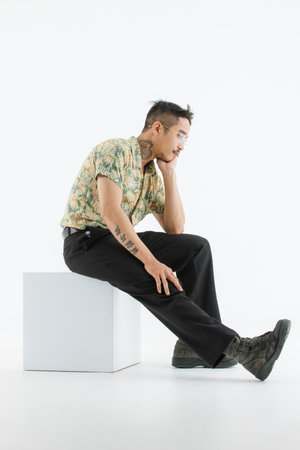 Isolated cutout studio shot Asian vintage classic male fashion model with mustache neck arm tattoo wears eyeglasses in casual classy outfit and leather boots sitting on square box on white background.の写真素材