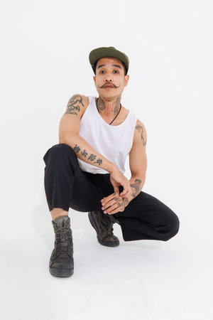 Isolated cutout studio shot Asian vintage classy mustache with neck arms hands tattoos male fashion model in casual fashionable sleeveless shirt cap boots sitting on floor on white background.の写真素材