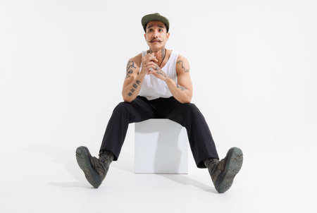 Isolated cutout studio shot Asian vintage classy mustache with neck arms hands tattoos male fashion model in casual fashionable sleeveless shirt cap boots sitting on square box on white background.の写真素材