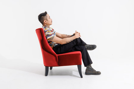 Portrait isolated cutout studio shot Asian vintage classy mustache neck arms hands tattoos male fashion model in casual fashionable classic outfit leather boots sitting on red chair white backgroundの写真素材