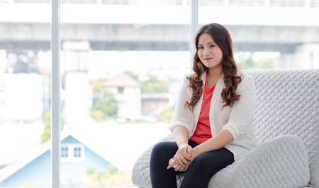 Asian young beautiful overweight oversized fat chubby plump professional successful businesswoman in casual outfit jacket sitting smiling looking at camera on cozy sofa couch in office living room.の写真素材