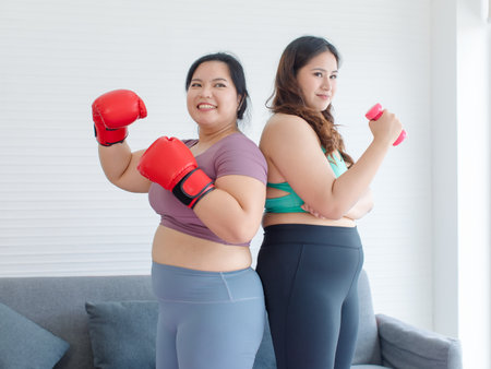 TWO Asian young chubby fat overweight fit female sportswoman in casual sportswear and boxing gloves standing posting together on yoga mat smiling punching air exercising training in home fitness gym.の写真素材