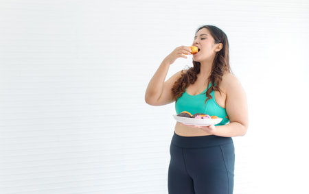 Asian young chubby fat unhealthy oversized overweight plus size female sportswoman in sportswear sports bra legging standing  holding eating mixed flavor sweet sugar dessert donuts from plate.の写真素材
