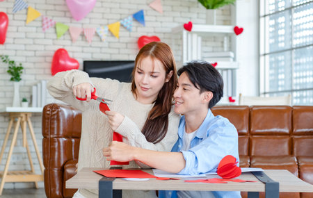 Asian young handsome male boyfriend sitting on sofa smiling cuddling with beautiful female girlfriend holding red heart shape paper cutting in romantic decorated living room celebrating valentine day.の写真素材