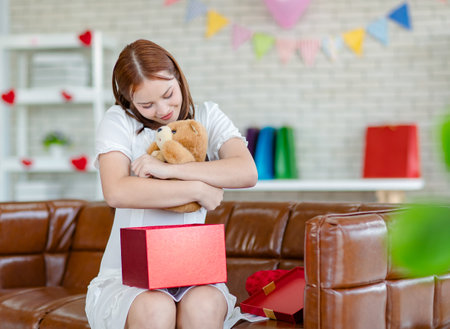 Asian beautiful female girlfriend in white dress sitting on cozy sofa in decorated living room celebrating valentine day opening unboxing wrapped gift present box holding hugging teddy bear doll.の写真素材