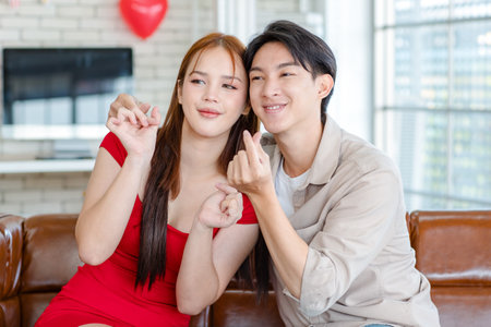 Asian young handsome male boyfriend and beautiful female girlfriend in red dress sitting smiling together on sofa posing hand heart shape  celebrating valentine day in living room at home.の写真素材