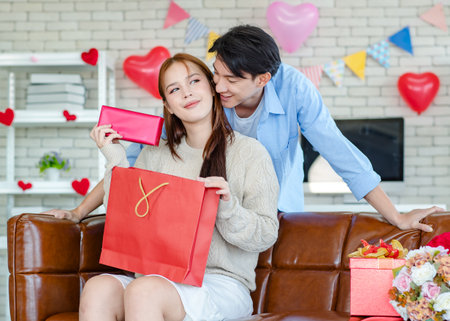 Asian young handsome male boyfriend standing smiling behind beautiful female girlfriend giving red wrapped present gift box in paper bag in romantic decorated living room celebrating valentine day.の写真素材