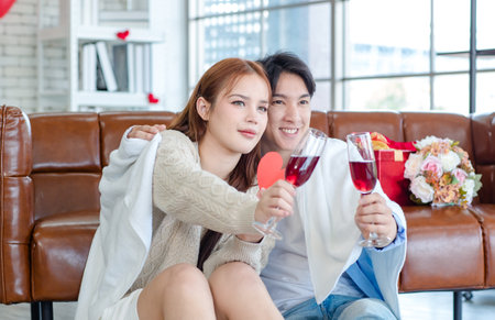 Asian young handsome male boyfriend sitting smiling cuddling together with beautiful female girlfriend holding tall wine glass cheers toasting in romantic decor living room celebrating valentine day.の写真素材