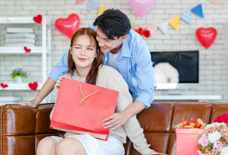 Asian young handsome male boyfriend standing smiling behind beautiful female girlfriend giving red wrapped present gift box in paper bag in romantic decorated living room celebrating valentine day.の写真素材