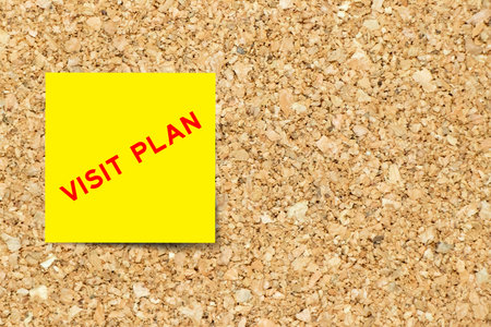 Yellow note paper with word visit plan on cork board background with copy space