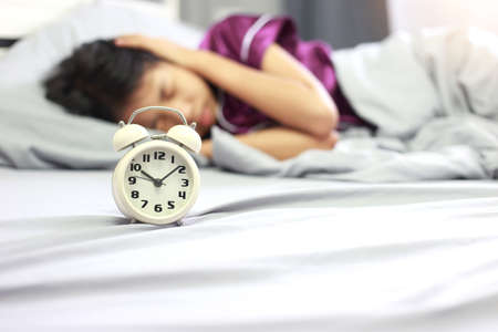 Wake up concept - Asian girl covering her ear with hand from alarm clock noise in her bedroom in the morning. Upset asian girl being awaken with alarm clock.の写真素材