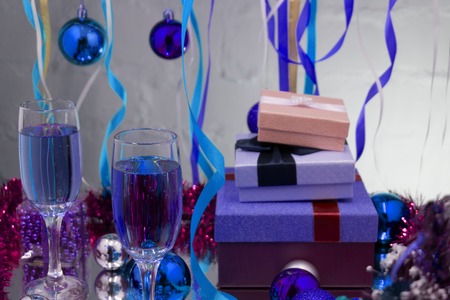 A couple of glasses with champagne on a wooden table with Christmas red balls, a glossy ribbon with a sprig of spruce on the background with a blue bokeh. Happy new year concept photo. Shallow DOFの写真素材